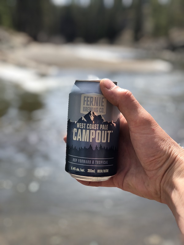 A hand holding a can of campout by a river