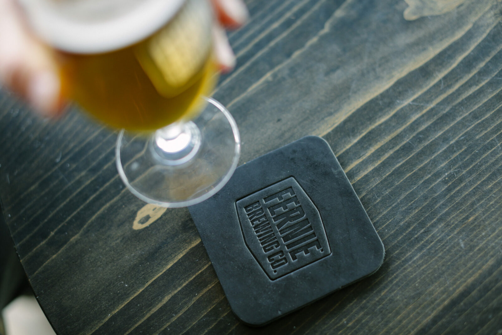 Glass of beer being lifted off of a Fernie Brewing Co. black leather coaster.