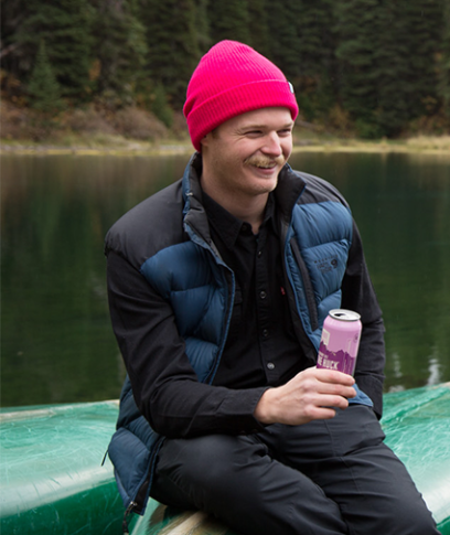 Man holding a What the Huck-Huckleberry Ale sat on a canoe next to a lake