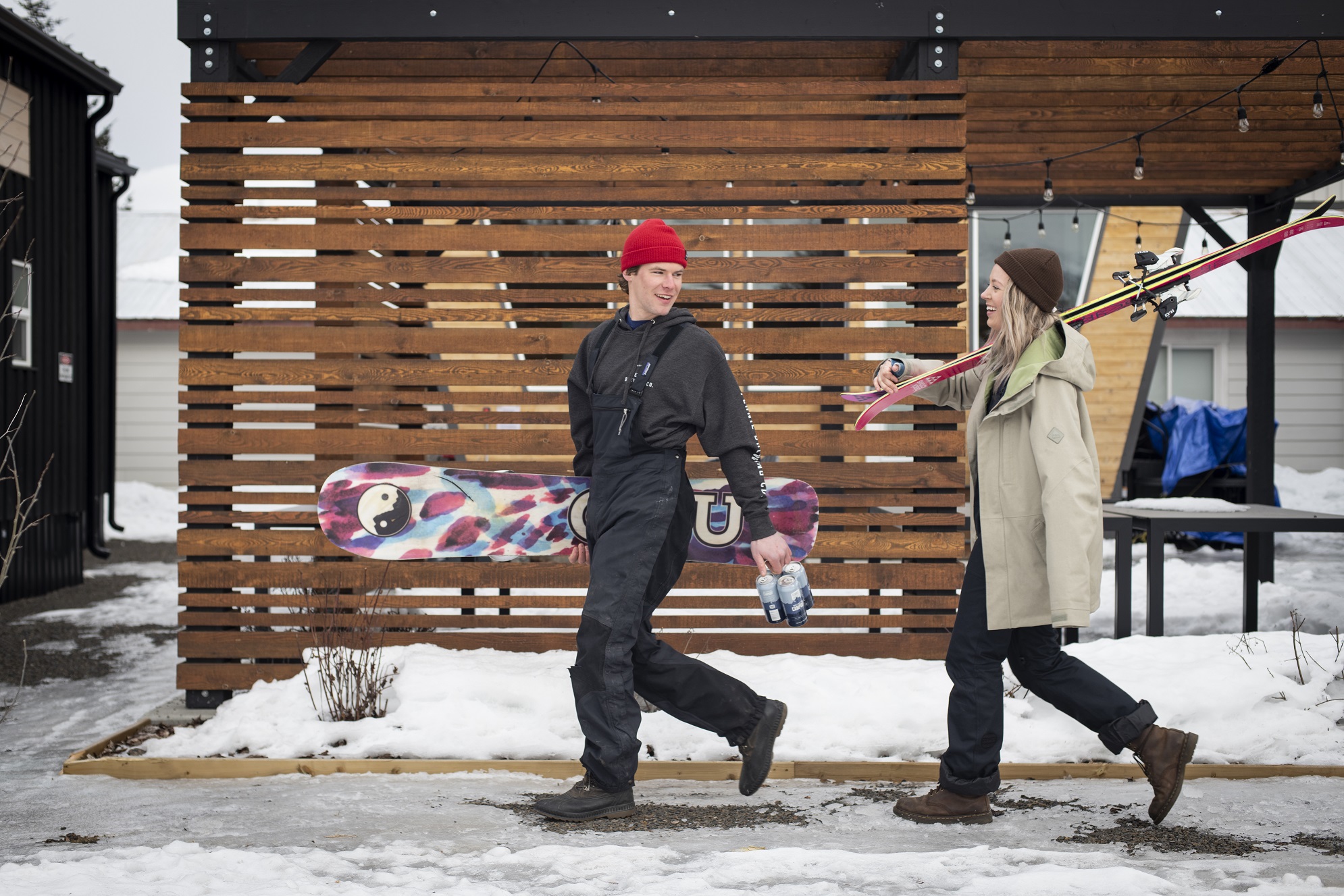 Man holding a snowboard and beers walking with a women holding skis during winter.