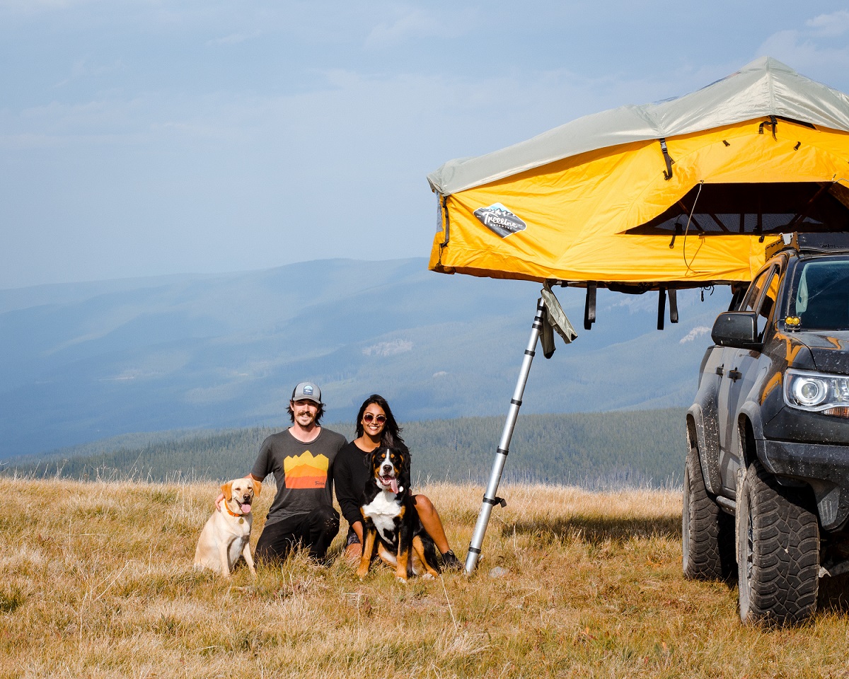 A man and woman kneel next to their 2 dogs and offroad truck in a field on a mountain top