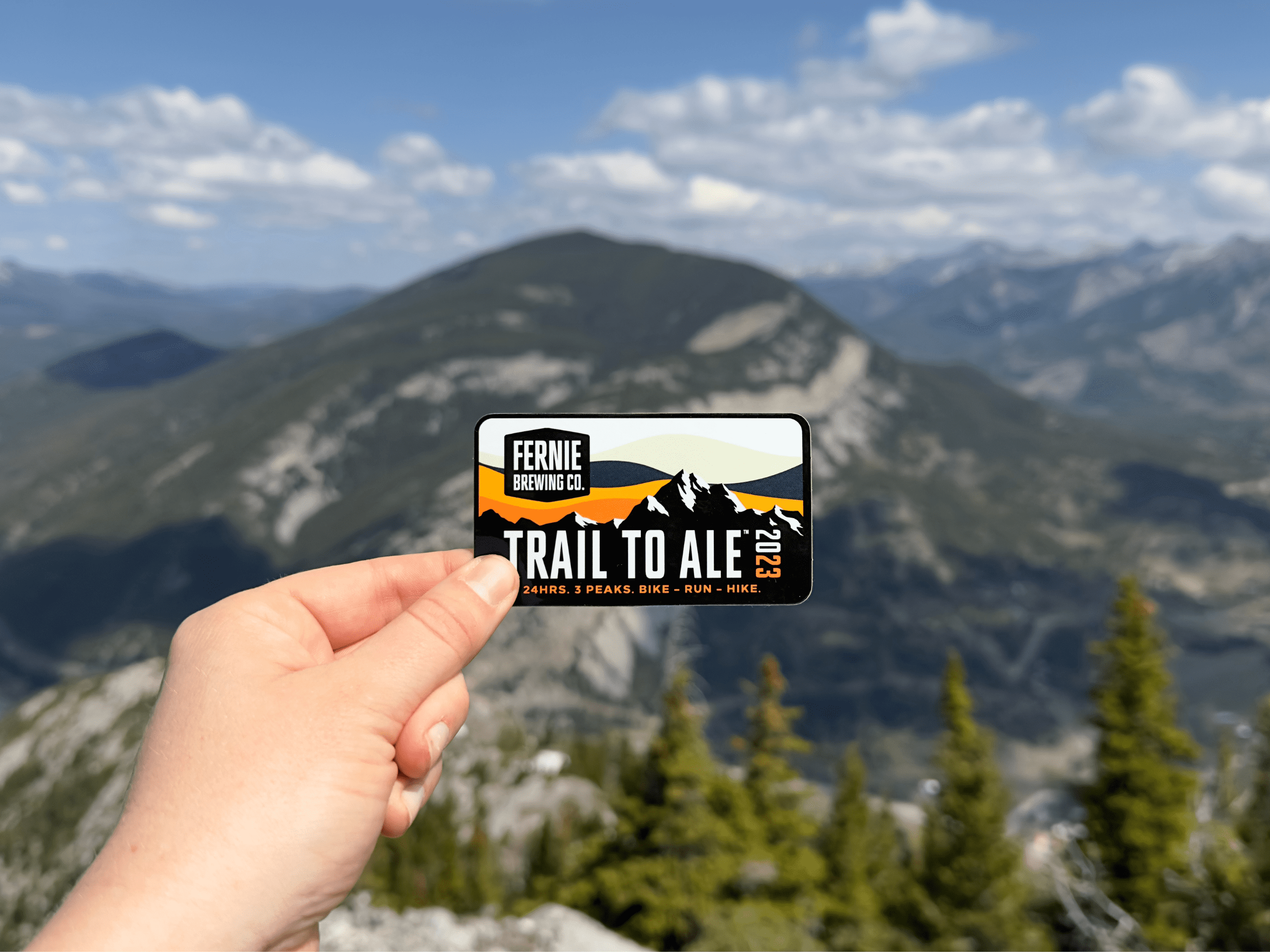 A person holding a Trail to Ale 2023 sticker with a mountain view in the background.